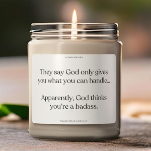 May include: A lit candle in a glass jar with a silver lid. The label reads, "They say God only gives you what you can handle... Apparently, God thinks you're a badass." The candle sits on a wooden surface.