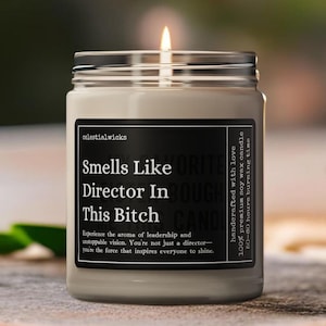 May include: A black and white candle jar with the text "Smells Like Director In This Bitch" and the text "Experience the aroma of leadership and unstoppable vision. You're not just a director - you're the force that inspires everyone to shine." The candle is lit and the jar is sitting on a wooden surface.