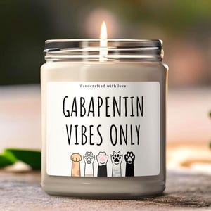 May include: A lit, grey candle in a glass jar with a silver lid. The label reads "GABAPENTIN VIBES ONLY" with paw prints. The words "handcrafted with love" are above the main text. The candle sits on a wooden surface.