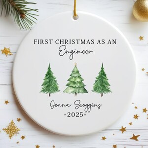 Op de afbeelding: Ronde witte keramische kerstversiering met een gouden ophangtouwtje. Het heeft de tekst "FIRST CHRISTMAS AS AN Engineer" en drie groene aquarel kerstbomen. De naam "Jenna Scogging" en het jaar "-2025" staan er ook op.