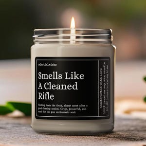 May include: A lit soy wax candle in a glass jar with a silver lid. The black label reads "Smells Like A Cleaned Rifle." The candle is on a wooden surface. The text on the label also says "handcrafted with love" and "50-60 hours burning time."