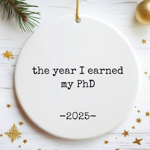 Peut inclure: Ornement de Noël rond en céramique blanche avec l'inscription "the year I earned my PhD -2025-". Il est suspendu à un cordon doré. Des étoiles et boules dorées décorent l'ornement, avec une branche de sapin.