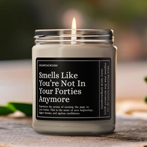 May include: A burning candle in a glass jar with a silver lid. The label says "Smells Like You're Not In Your Forties Anymore." The candle sits on a wooden surface. The label also states "handcrafted with love" and "100% premium soy wax candle".