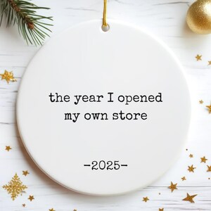 May include: A white, round Christmas ornament with the text "the year I opened my own store -2025-". The ornament is hanging by a gold string, with gold star and ball decorations, and evergreen branches in the background.