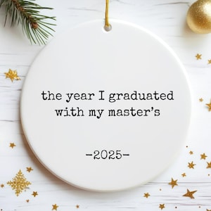 Può includere: Un ornamento rotondo in ceramica bianca con la scritta "the year I graduated with my master's -2025-". L'ornamento è sospeso da un filo d'oro e circondato da stelle dorate, ornamenti e rami di sempreverdi.