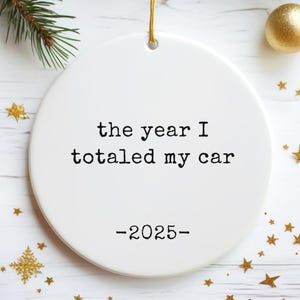May include: A white, round Christmas ornament with the text "the year I totaled my car -2025-" in black. The ornament hangs from a gold string. Gold star and ball ornaments and pine branches are in the background.