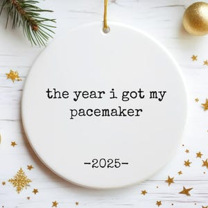May include: White ceramic Christmas ornament with the text "the year i got my pacemaker -2025-". The ornament is round and has a gold string for hanging. Gold star and ball ornaments and pine branches surround the ornament.