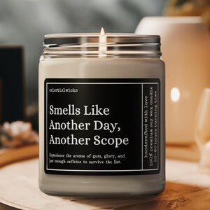 May include: A lit soy wax candle in a glass jar with a silver lid. The label reads "Smells Like Another Day, Another Scope." The candle is on a wooden tray, with a blurred background. The text "Celestial Giftsz" is in the top right corner.