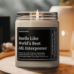 May include: A lit soy wax candle in a glass jar with a silver lid. The label reads "Smells Like World's Best ASL Interpreter." The candle is handcrafted with love and has a 50-60 hour burn time. The brand name "CelestialWicks" is visible.