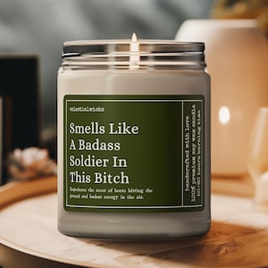 May include: A glass jar candle with a silver lid and a white label. The label reads "Smells Like A Badass Soldier In This Bitch." The candle is on a wooden tray with other decorative items. The candle is handcrafted with love and made of 100% premium soy wax.
