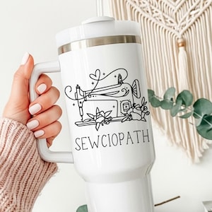 May include: White travel mug with a black illustration of a sewing machine, flowers, and the word "SEWCIOPATH". The mug has a white handle and lid. The mug is held by a person wearing a pink sweater.
