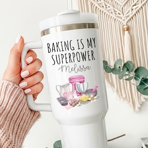 May include: White travel mug with a handle, featuring the text "BAKING IS MY SUPERPOWER Melissa" in black script. The mug has a lid and a graphic of baking tools and ingredients. The mug is held by a person wearing a pink sweater.
