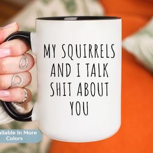 Op de afbeelding: Witte keramische mok met een zwarte binnenkant en handvat. De mok heeft de tekst "MY SQUIRRELS AND I TALK SHIT ABOUT YOU" in zwarte, vette letters. De mok wordt vastgehouden door een persoon met meerdere ringen aan hun vingers. De woorden "Available In More Colors" staan onderaan.