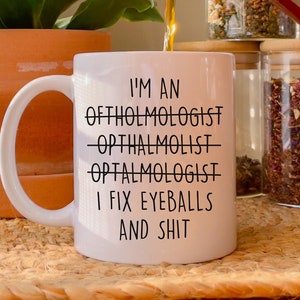 Może przedstawiać: Biały ceramiczny kubek do kawy z czarnym tekstem, który brzmi "I'm an Ophtholmologist -Opthalmolist- Optalmologist I fix eyeballs and shit". Kubek jest napełniany brązową cieczą.