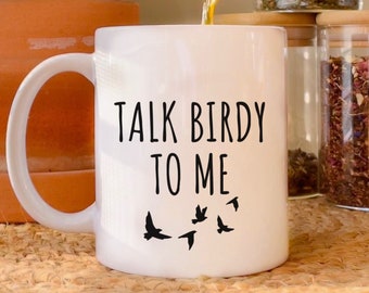 Op de afbeelding: Een witte keramische mok met zwarte tekst die "Talk Birdy To Me" zegt en een ontwerp van zwarte vogels die vliegen.