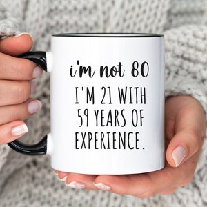 May include: White ceramic mug with a black rim and handle. The mug has black text that reads "i'm not 80 I'm 21 with 59 years of experience."