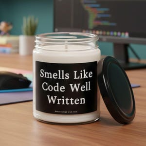 May include: A white candle in a clear glass jar with a black lid. The candle has a black label with the text "Smells Like Code Well Written". The candle sits on a wooden desk next to a computer mouse and keyboard.