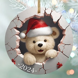 May include: A round Christmas ornament featuring a teddy bear wearing a Santa hat, peeking through a cracked surface. The ornament is white with a gold string for hanging. The year "2024" is printed on the ornament.