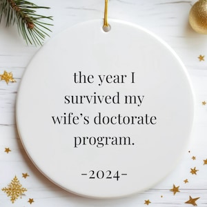 Puede incluir: Adorno navideño redondo de cerámica blanca con cordón dorado. El texto dice: "the year I survived my wife's doctorate program. -2024-". Estrellas doradas, bolas y ramas de pino en el fondo.