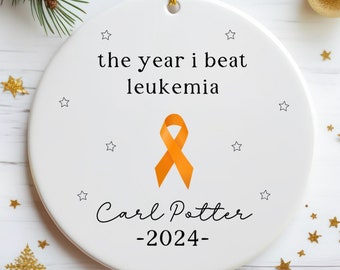May include: White ceramic Christmas ornament with an orange ribbon, the text "the year i beat leukemia", the name "Carl Potter", and the year "2024".