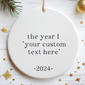 May include: White ceramic Christmas bauble with the text "the year I *your custom text here* -2024- ". The bauble is surrounded by gold glitter stars on a white wooden background.