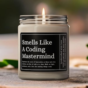 May include: A lit, grey candle in a glass jar with a black label that reads "Smells Like A Coding Mastermind." The candle is on a wooden surface. The label also states the candle is handcrafted with love and made of 100% premium soy wax.
