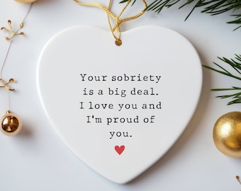 May include: A white heart-shaped ceramic ornament featuring the black printed text: Your sobriety is a big deal. I love you and I'm proud of you. A small red heart is printed below the text. The ornament hangs from a thin gold string.