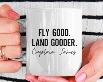 以下が含まれることがあります： 白いセラミック製のコーヒーカップで、黒い文字で「Fly good. Land gooder. Captain James」と書かれています。