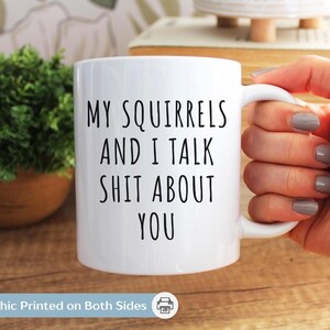 May include: White ceramic mug with the text "MY SQUIRRELS AND I TALK SHIT ABOUT YOU" in black. The mug has a white handle and is held by a person. A small green plant in a brown pot is in the background.