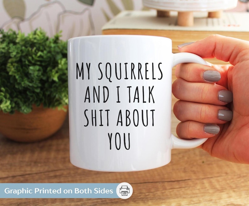 May include: White ceramic mug with the text "MY SQUIRRELS AND I TALK SHIT ABOUT YOU" in black. The mug has a white handle and is held by a person. A small green plant in a brown pot is in the background.