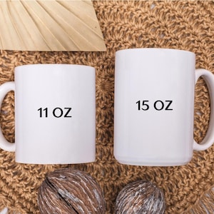 May include: Two white ceramic mugs, one labeled "11 OZ" and the other "15 OZ". The mugs are displayed on a textured, brown surface. The mugs have large handles and a cylindrical shape.