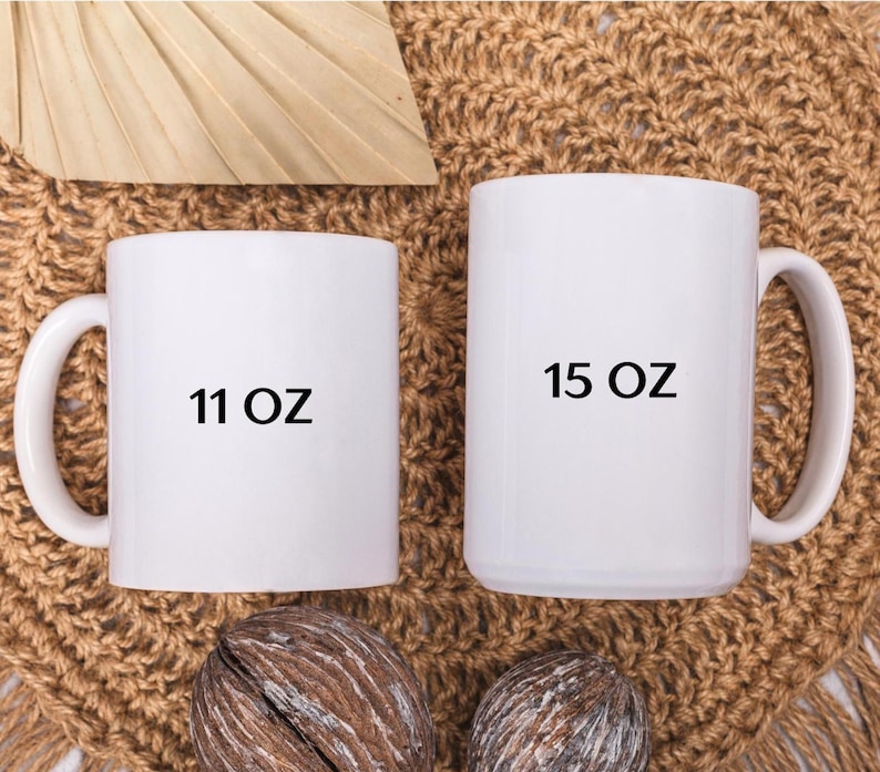 May include: Two white ceramic mugs, one labeled "11 OZ" and the other "15 OZ". The mugs are displayed on a textured, brown surface. The mugs have large handles and a cylindrical shape.