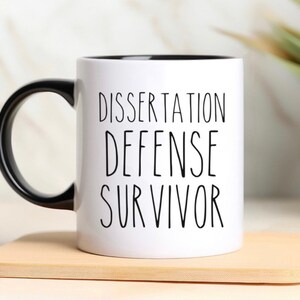 May include: White ceramic coffee mug with a black handle and black rim. The mug has the text "Dissertation Defense Survivor" printed in black.