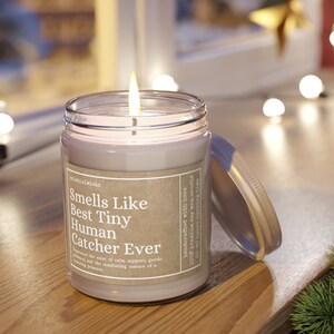 May include: A clear glass candle jar with a brown label that reads "Smells Like Best Tiny Human Catcher Ever". The candle is lit and the label also says "Handcrafted with love, 100% premium soy wax candle, 50-80 hours burning time".