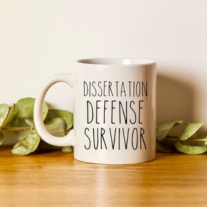 May include: White ceramic coffee mug with black text that reads "Dissertation Defense Survivor".