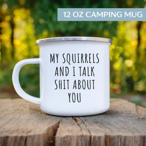 May include: White 12 oz camping mug with a silver rim and a large white handle. The mug has the text "MY SQUIRRELS AND I TALK SHIT ABOUT YOU" in black. The mug is sitting on a wooden surface.