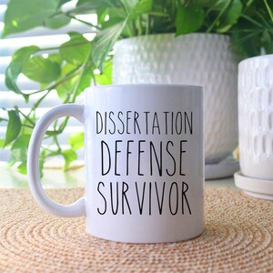 May include: White ceramic coffee mug with black text that reads "Dissertation Defense Survivor".