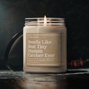 May include: A brown glass candle jar with a beige label that reads "Smells Like Best Tiny Human Catcher Ever". The label also includes the text "Experience the scent of calm support, gentle guidance, and the comforting essence of a nurturing presence." The candle is lit and the flame is visible.