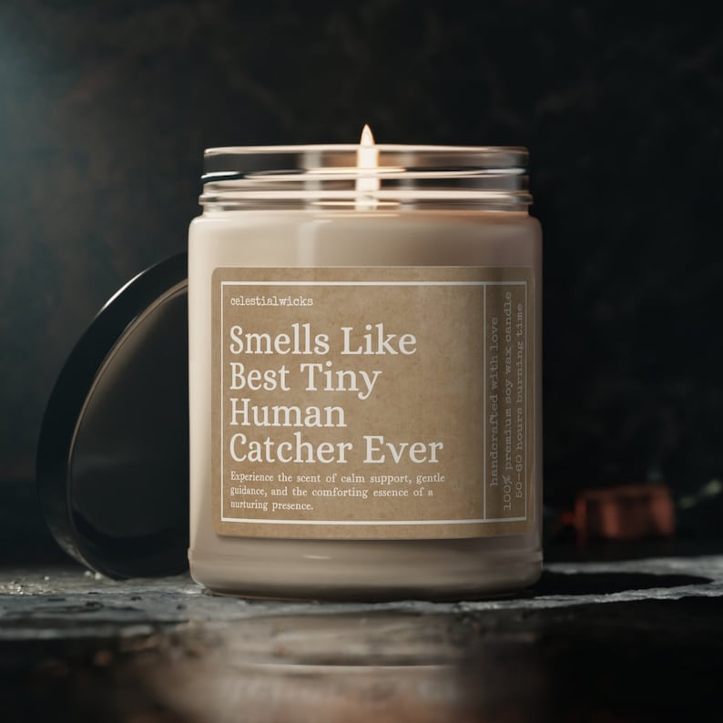 May include: A brown glass candle jar with a beige label that reads "Smells Like Best Tiny Human Catcher Ever". The label also includes the text "Experience the scent of calm support, gentle guidance, and the comforting essence of a nurturing presence." The candle is lit and the flame is visible.