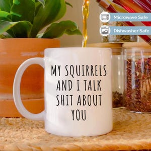 May include: White ceramic mug with a large handle, featuring the text "MY SQUIRRELS AND I TALK SHIT ABOUT YOU" in black. The mug is near a potted plant and glass jars. The mug is microwave and dishwasher safe.