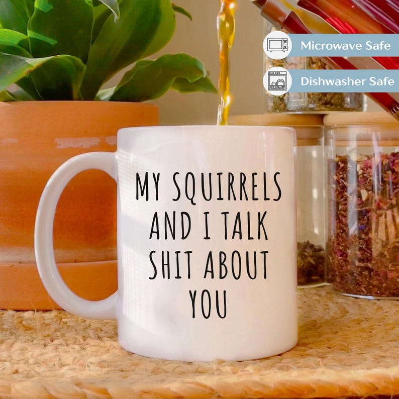 May include: White ceramic mug with a large handle, featuring the text "MY SQUIRRELS AND I TALK SHIT ABOUT YOU" in black. The mug is near a potted plant and glass jars. The mug is microwave and dishwasher safe.