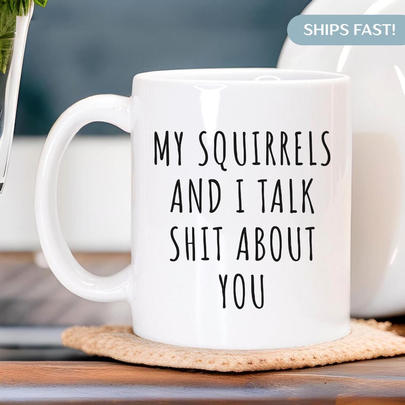 May include: White ceramic mug with the text "MY SQUIRRELS AND I TALK SHIT ABOUT YOU" in black, with a curved handle. The mug is sitting on a tan coaster.