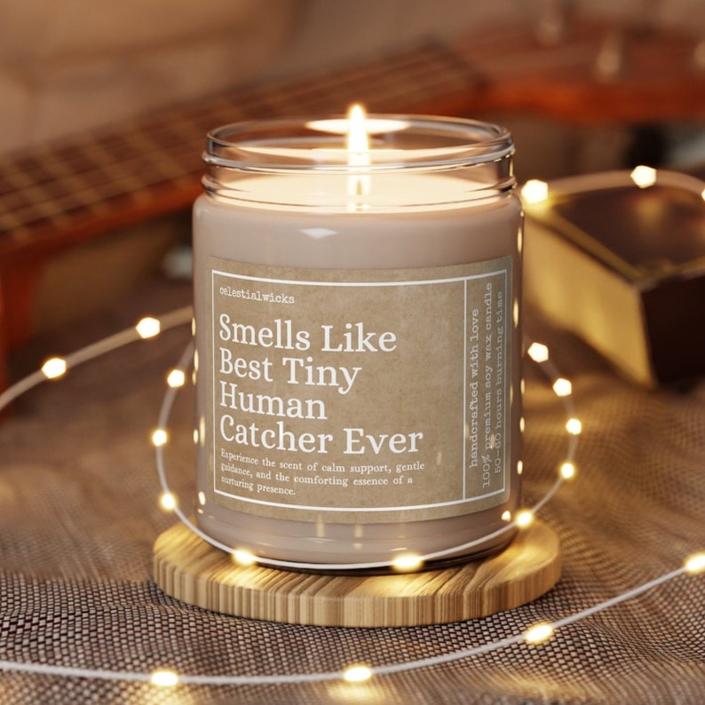 May include: A brown soy wax candle in a clear glass jar with a label that reads "Smells Like Best Tiny Human Catcher Ever". The candle is sitting on a wooden coaster and surrounded by white string lights.
