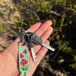 May include: A green woven keychain with a red mushroom and pink flower design, attached to a silver clasp and two keys. The keychain has a tassel at the bottom. The background is blurred.