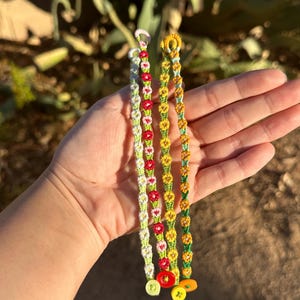 May include: Three friendship bracelets with flower patterns in green, white, red, and yellow. The bracelets are tied with colorful buttons.
