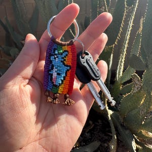 May include: A colorful handmade keychain with a rainbow design and a blue bear figure. The keychain is attached to a silver key ring with two keys. The keychain has small brown tassels at the bottom.