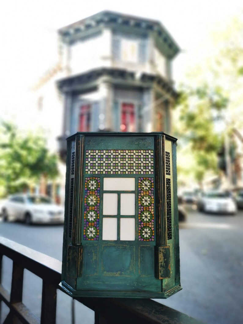 K&ouml;nnte beinhalten: Eine gr&uuml;ne, h&ouml;lzerne Dekorationsbox mit einem Buntglasfenster. Das Fenster hat ein geometrisches Muster mit bunten Glasscheiben. Die Box steht auf einem schwarzen Metallgel&auml;nder.