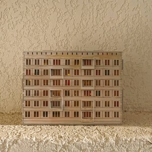 May include: A miniature model of a beige apartment building with multiple windows. The windows are lit with small orange lights, giving the impression of a building at night.