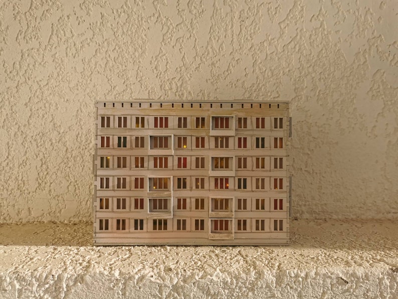 May include: A miniature model of a beige apartment building with multiple windows. The windows are lit with small orange lights, giving the impression of a building at night.