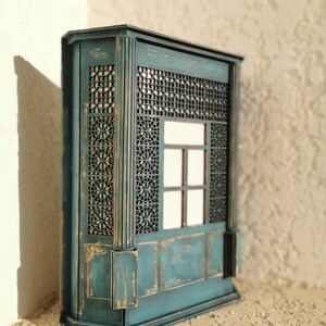 K&ouml;nnte beinhalten: Ein blaugr&uuml;ner Holzschrank mit einem dekorativen Gitterfenster und einer abgenutzten Oberfl&auml;che. Der Schrank hat ein kleines Regal und eine get&auml;felte Unterseite.